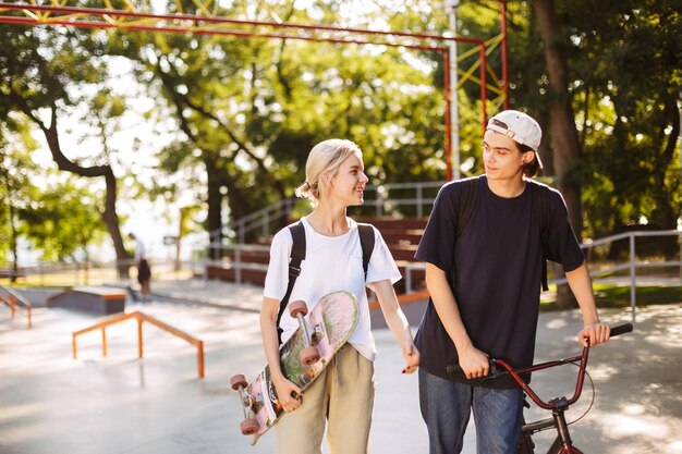 Young guy with bicycle and pretty smiling girl with skateboard joyfully spending time together at modern skatepark