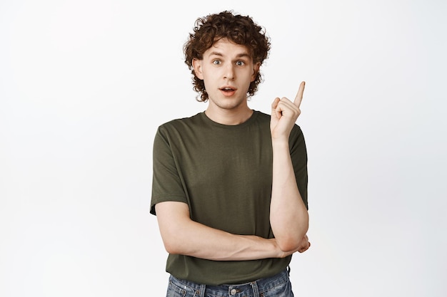 Young guy with curly natural hair pointing finger up raising eyebrows up surprised showing advertisement standing over white background