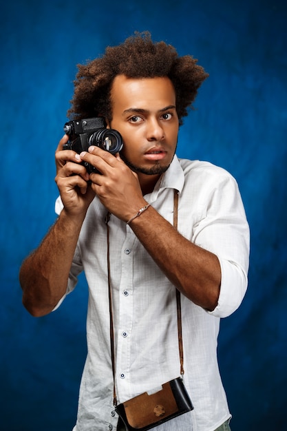 Young handsome african man holding old camera over blue wall.