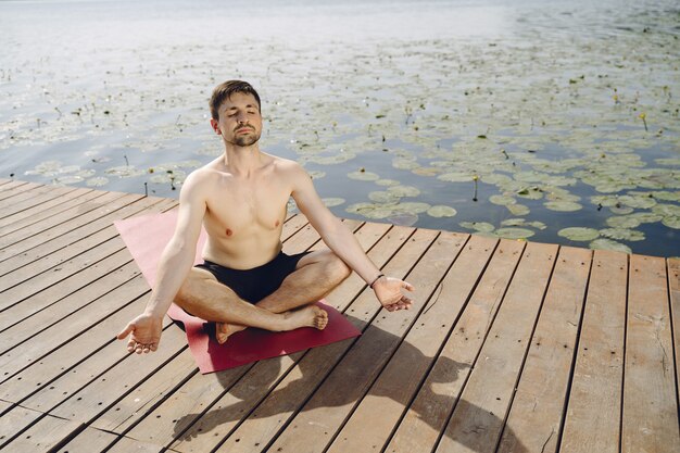 Young handsome bearded man sitting on wooden pier in summer day. Meditating and relaxing.