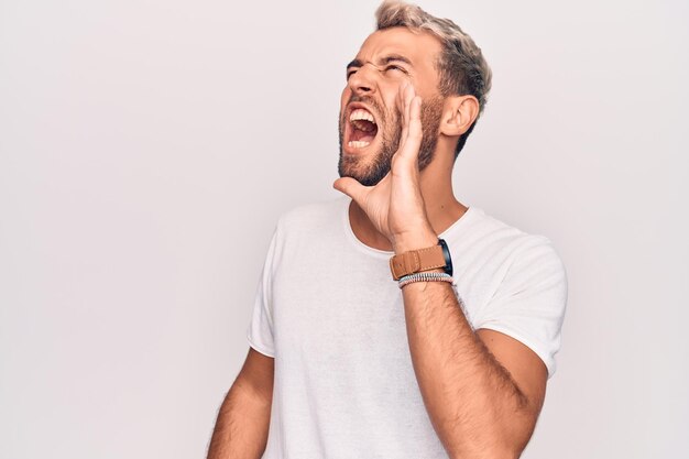 Young handsome blond man wearing casual tshirt standing over isolated white background shouting and screaming loud to side with hand on mouth Communication concept