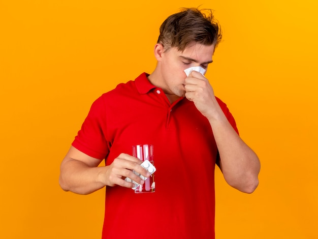 Young handsome blonde ill man holding pack of medical tablets and glass of water wiping nose with napkin with closed eyes isolated on orange background