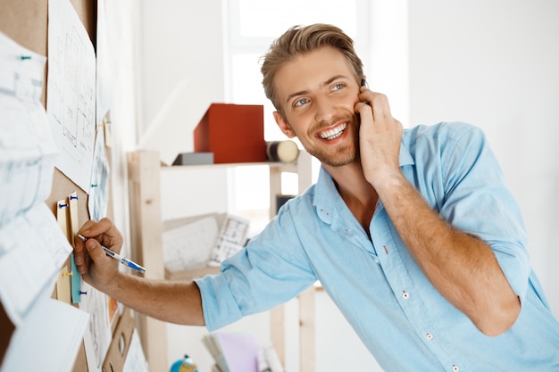 Free photo young handsome businessman writing at the paper pinned to corkboard, talking on  phone smiling.