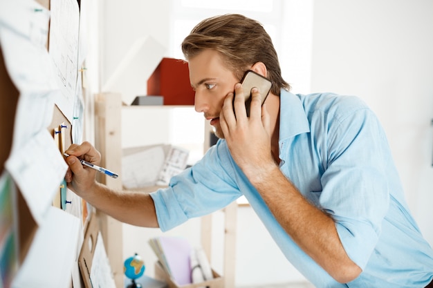 Young handsome businessman writing at the paper pinned to corkboard, talking on  phone.