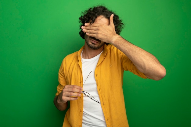 Free photo young handsome caucasian man holding glasses covering eyes with hand isolated on green background with copy space