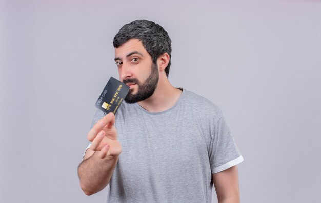 Young handsome caucasian man stretching out credit card towards camera and looking at it isolated on white background with copy space