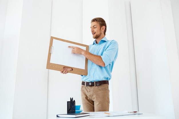 Free photo young handsome cheerful businessman standing at table holding wooden clipboard with white sheet pointing on it. light modern office interior