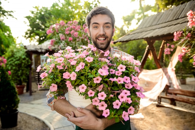 Young handsome cheerful gardener smiling, holding big pot with flowers