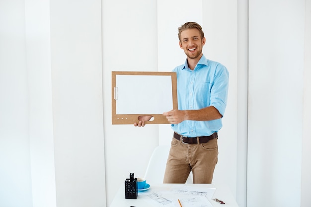 Free photo young handsome cheerful smiling businessman standing at table holding wooden clipboard with white sheet. light modern office interior