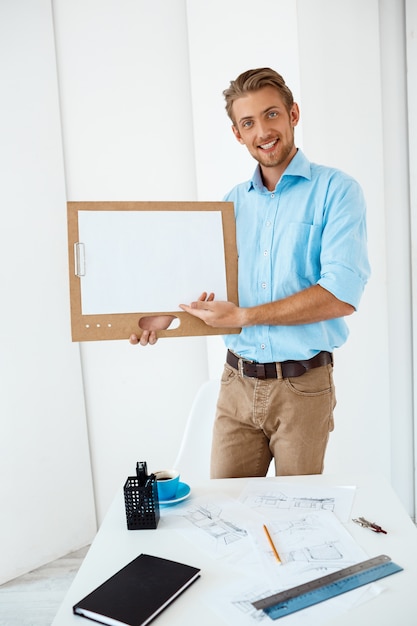 Free photo young handsome cheerful smiling businessman standing at table holding wooden clipboard with white sheet pointing on it. light modern office interior