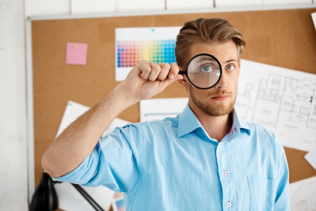 Free photo young handsome confident pensive businessman standing over cork board  through magnifier. white modern office interior