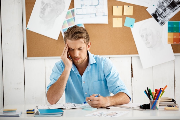 Free photo young handsome confident pensive businessman working sitting at table thinking over notepad. white modern office interior