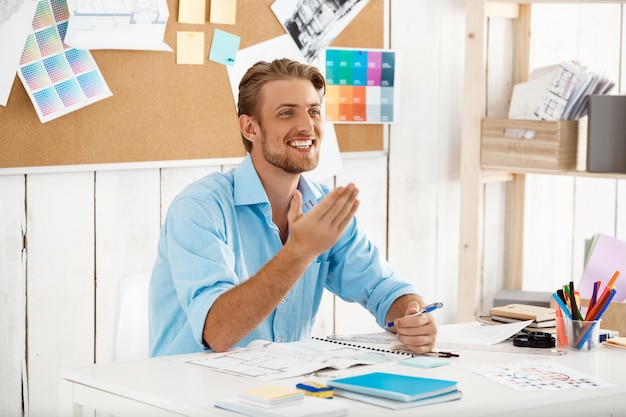 Young handsome confident smiling businessman working sitting at table. White modern office interior 