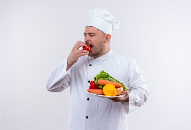 Free photo young handsome cook in chef uniform holding plate with vegetables and biting tomato with closed eyes on isolated white space