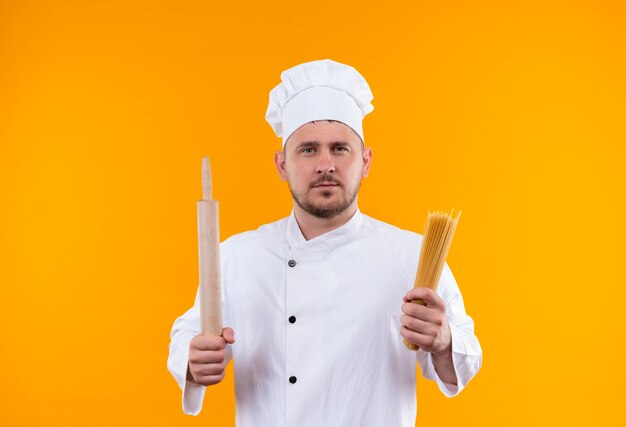 Young handsome cook in chef uniform holding rolling pin and spaghetti pasta looking  isolated on orange space