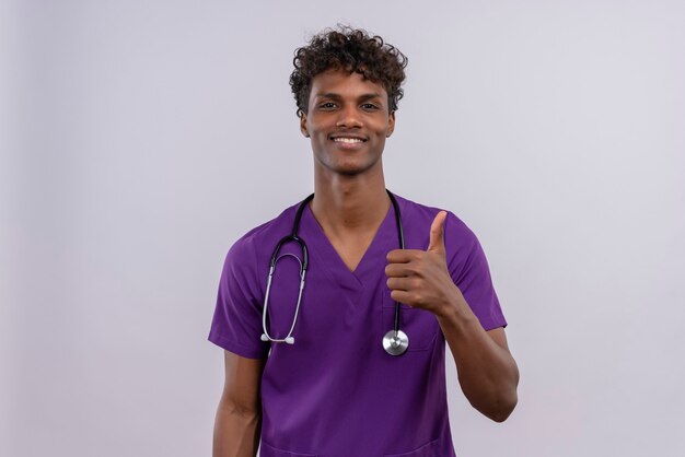 A young handsome dark-skinned doctor with curly hair wearing violet uniform with stethoscope happy while showing thumbs up 