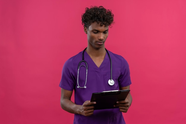 A young handsome dark-skinned doctor with curly hair wearing violet uniform with stethoscope looking at clipboard 