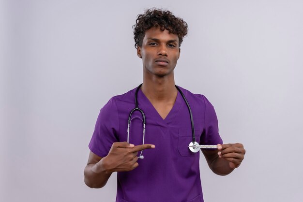 A young handsome dark-skinned doctor with curly hair wearing violet uniform with stethoscope showing thumbs down while pointing at thermometer with index finger 