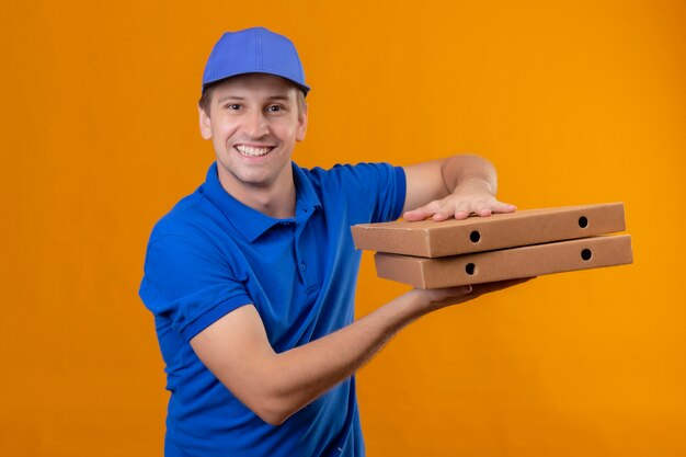 Young handsome delivery man in blue uniform and cap holding pizza boxes smiling friendly standing over orange wall