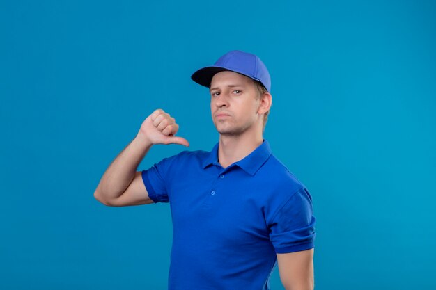 Young handsome delivery man in blue uniform and cap pointing to himself with thumb looking confident self-satisfied and proud standing over blue wall