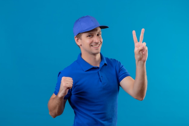 Young handsome delivery man in blue uniform and cap showing number two smiling cheerfully standing over blue wall