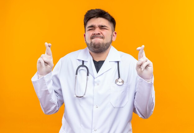 Young handsome doctor wearing white medical gown white medical gloves and stethoscope closing eyes