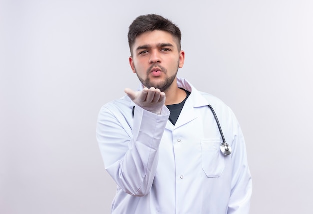 Young handsome doctor wearing white medical gown white medical gloves and stethoscope sending kisses standing over white wall