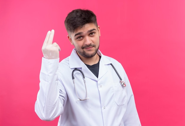 Young handsome doctor wearing white medical gown white medical gloves and stethoscope with cool face showing two fingers up standing over pink wall