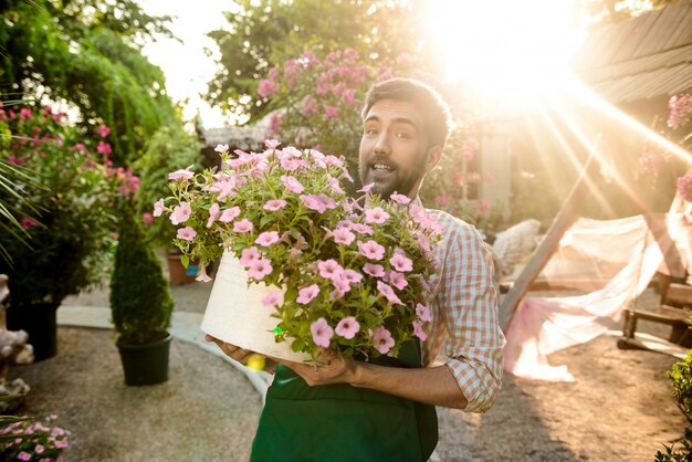 Young handsome gardener smiling, holding big pot with flowers