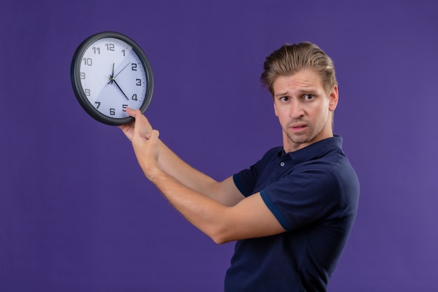 Young handsome guy holding clock looking at camera with confuse expression standing over purple background