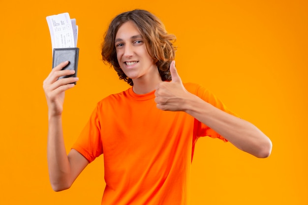 Young handsome guy in orange t-shirt holding air tickets looking confident showing thumbs up smiling cheerfully standing