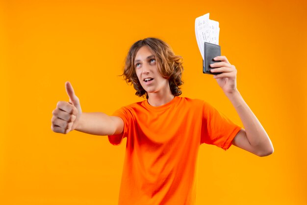 Young handsome guy in orange t-shirt holding air tickets looking confident showing thumbs up smiling cheerfully standing