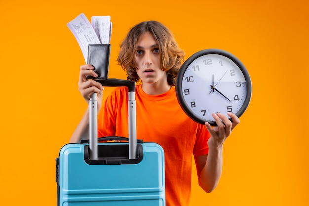 Free photo young handsome guy in orange t-shirt holding travel suitcase and air tickets standing with clock looking confused