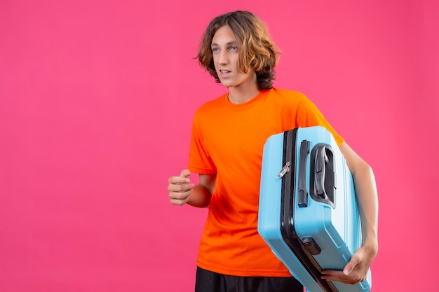 Free Photo young handsome guy in orange t-shirt holding travel suitcase looking aside smiling standing over pink background