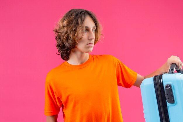 Young handsome guy in orange t-shirt holding travel suitcase looking aside with serious face standing