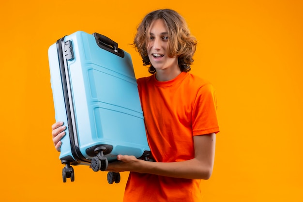 Free photo young handsome guy in orange t-shirt holding travel suitcase positive and happy smiling cheerfully standing over yellow background