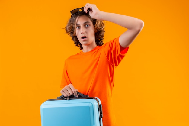 Free photo young handsome guy in orange t-shirt holding travel suitcase putting off his glasses looking surprised and amazed standing