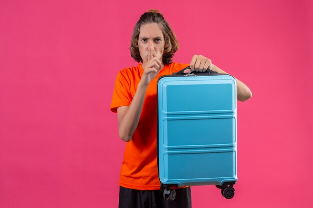 Young handsome guy in orange t-shirt standing with travel suitcase making silence gesture with finger on lips