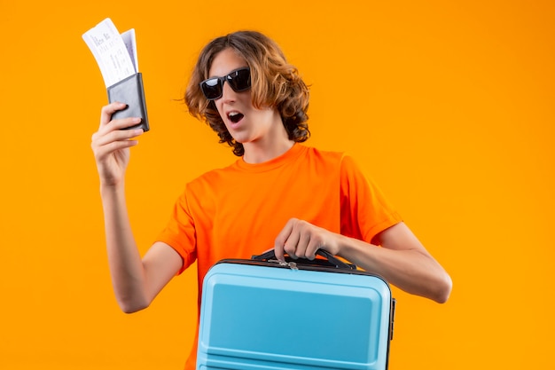 Free Photo young handsome guy in orange t-shirt wearing black sunglasses holding air tickets and travel suitcase looking surprised and happy standing