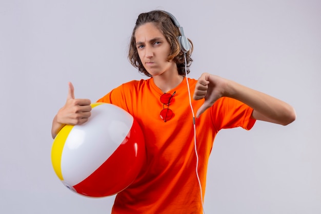 Free photo young handsome guy in orange t-shirt with headphones holding inflatable ball displeased showing thumbs up and down standing over white background