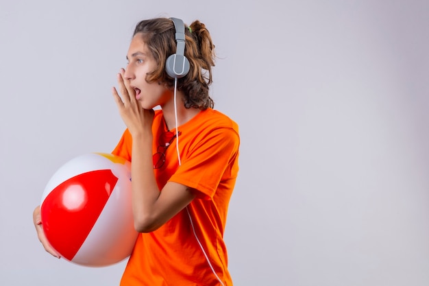 Free Photo young handsome guy in orange t-shirt with headphones holding inflatable ball looking aside telling a secret with hand near mouth standing over white background