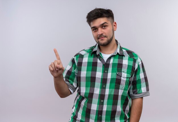 Young handsome guy wearing checkered shirt smiling pointing up with forefinger standing over white wall