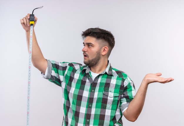 Free Photo young handsome guy wearing checkered shirt surprised holding measurer standing over white wall