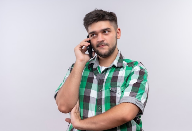 Young handsome guy wearing checkered shirt talking on phone looking besides standing over white wall