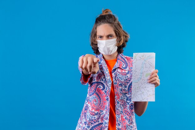 Young handsome guy wearing facial protective mask showing map pointing to camera angry and furious with you standing