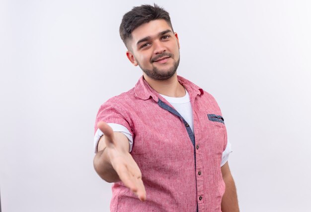 Young handsome guy wearing pink polo shirt coridally giving hand for handshaking standing over white wall