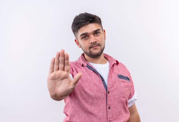 Young handsome guy wearing pink polo shirt doing stop sign with hand standing over white wall