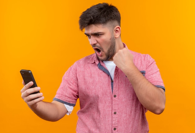 Young handsome guy wearing pink polo shirt looking on phone doing fist up standing over orange wall