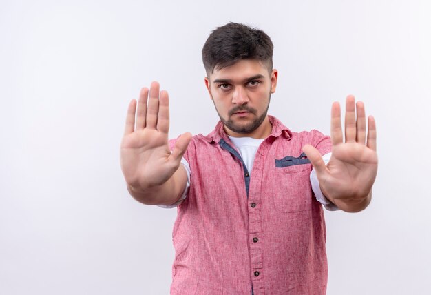 Young handsome guy wearing pink polo shirt looking seriously doing stop sign with hands standing over white wall