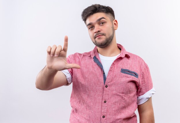 Young handsome guy wearing pink polo shirt looking seriously showing looser sign with hand standing over white wall